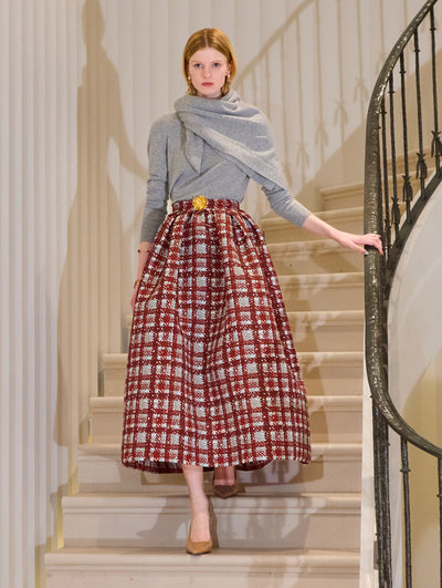 The model is wearing a red and blue plaid maxi skirt with a grey pullover and matching scarf. The look is styled with a blue belt and red sandals.  