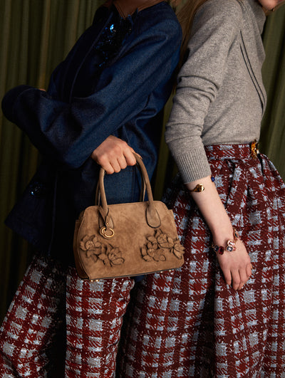 The model on the left is wearing a dark denim jacket and plaid pants. The styles feature a floral embroidery suede purse. The model is wearing a red and blue plaid maxi skirt with a grey pullover and matching scarf. The look is styled with a blue belt and red sandals.  