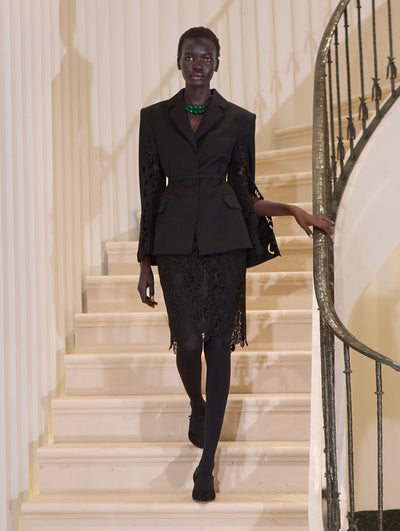 The model wears a lace guipure black blazer paired with a black lace skirt. The look is styled with black tights, a statement necklace and a clutch.  