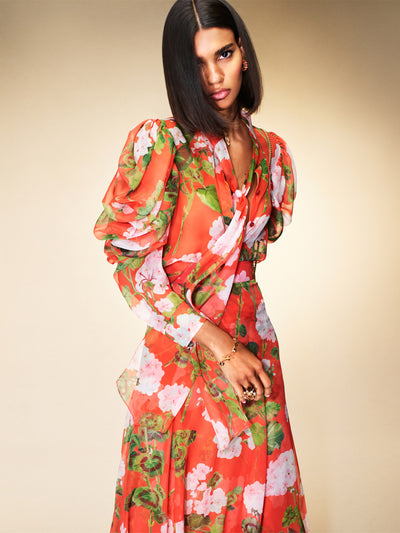 The model is wearing a red dress with large pink and white flowers. The dress has rouching in the sleeves and a necktie. 