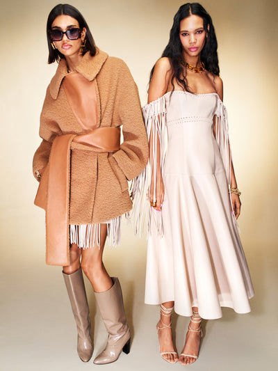 Two models pose next to one another. The model on the right is wearing a white off the shoulder dress with fringe tassels. The model on the left is wearing a brown teddy coat with a white fringe skirt. 