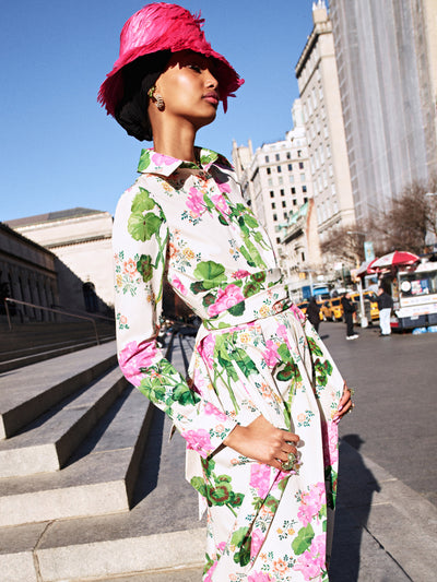 The model is wearing a long sleeve white dress with pink flowers and green leaves throughout with a pink feathered hat. 