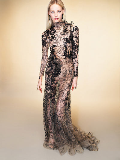 The model is wearing a sheer long sleeve gown with black floral applique. 