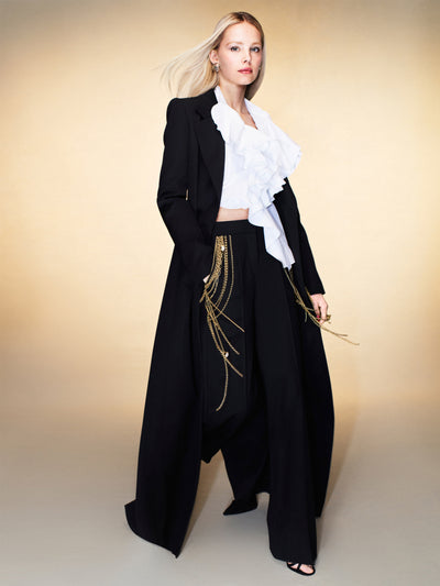The model is wearing black pants with gold fringe, a white blouse and a long black coat. 