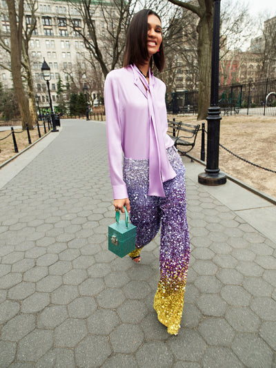 The model is wearing a lilac silk blouse and silver and purple sequin ombre pants. 