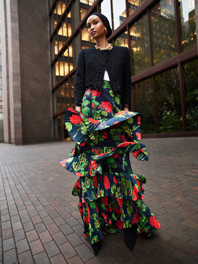 The model is wearing a black tweed jacket and a navy evening skirt with large red flowers and tiers of ruffle detailing. 