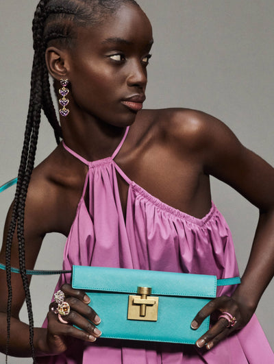A model poses against a grey back drop in an Oscar de la Renta purple dress with a bright blue handbag. 