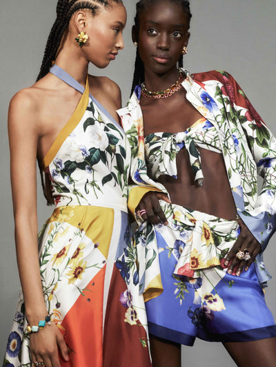 Two models pose against a grey backdrop in silk Oscar de la Renta ready-to-wear looks. One model is wearing an abstract floral handkerchief midi dress. The other model is wearing a floral blouse, bralette and short set. 