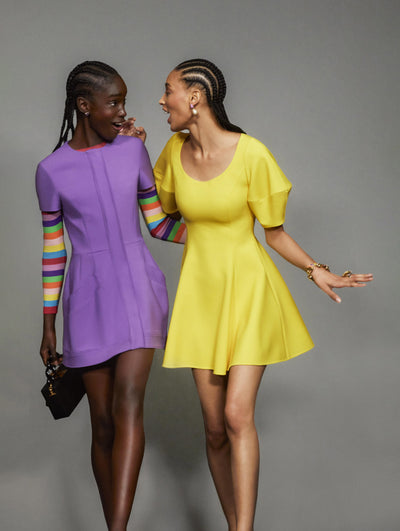 Two models pose against a grey backdrop in Oscar de la Renta ready-to-wear looks, one is wearing a purple day dress and the other is wearing a yellow day dress. 