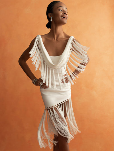The model is wearing a white mini dress with exaggerated fringe detailing at the hems. 