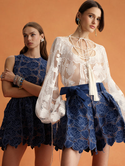 Two models pose next to each other. The model on the left is wearing a navy lace mini dress. The model on the right is wearing a white lace blouse and navy lace mini skirt. 