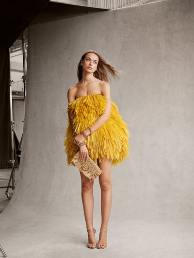The model is wearing a yellow strapless mini dress with fringe detailing. She is carrying a rattan clutch with a gold O clasp. 