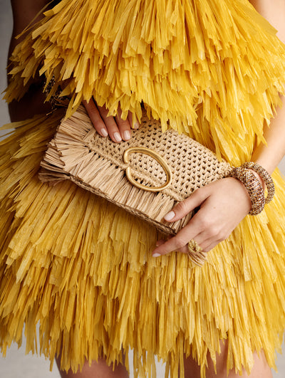 The model is wearing a yellow fringe mini dress and carrying a rattan clutch with a gold O clasp. 