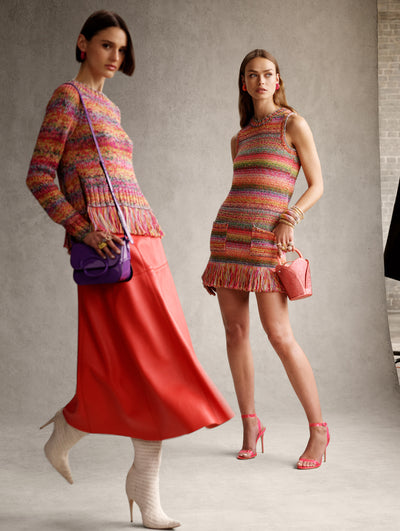 Two models pose next to each other. The model on the right is wearing a rainbow crochet mini dress. The model on the left is wearing a rainbow crochet sweater and orange maxi skirt. 