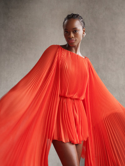 The model is wearing an orange mini dress with pleating and exaggerated sleeves. 