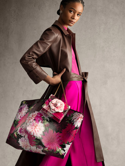 The model is wearing a chocolate brown leather coat and bright pink pants. She is carrying two pink floral purses. 