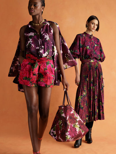 Two models pose next to each other. The model on the right is wearing a navy skirt and top set adorned with maroon flowers. The model on the left is wearing navy shorts with bright pink flowers and a pink top with maroon flowers. 