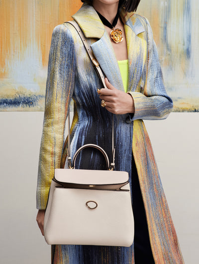 Model stands in front of paint backdrop wearing an abstract multicolor brushstroke trench coat with the Poppy handbag in ivory.