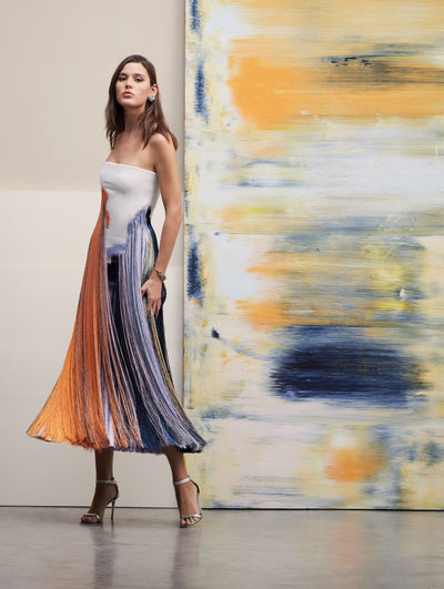 The model stands in front of a big brushstrokes painting wearing a strapless dress with abstract colors and fringe in tones of purple and orange.