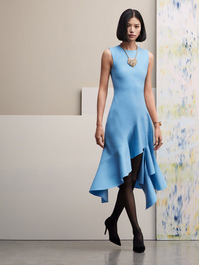The model wears a blue sleeveless dress, with a slit and ruffles in the bottom. It is paired with a heart necklace and black heels. Front View