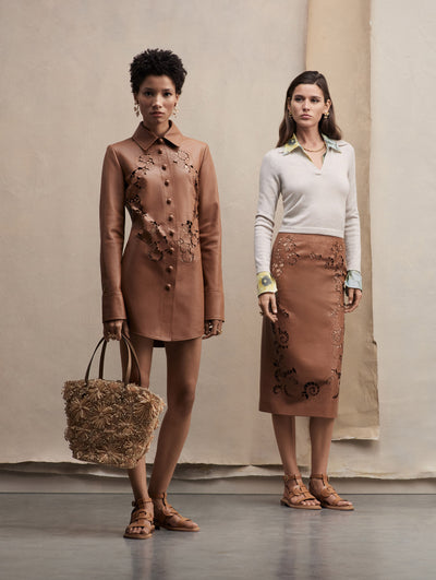 Two models pose together the model in the left wears a button-down leather mini dress with cutout details in brown, paired with a raffia tote bag and brown sandals. The model in the write wears a pencil skirt in the same brown with cutout details, paired with a polo collar knit.