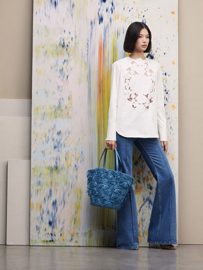 The model stands in front of a colorful painting while wearing a white blouse with floral cutouts in the middle, paired with jeans and a blue raffia flower tote.