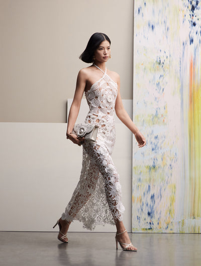 The model walks in a white crochet halter dress, while holding a white TRO handbag.