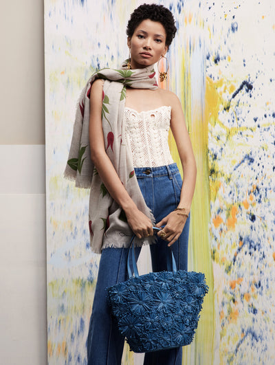 Model stands in front of splash paint backdrop wearing a white lace corset with jeans and a floral scarf, while holding a blue floral raffia tote.