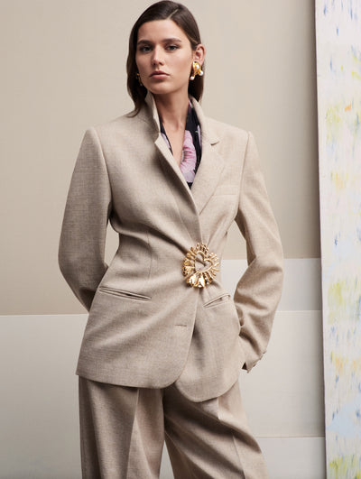 The model wears a beige blazer closed with a heart brooch and paired with matching beige pants.