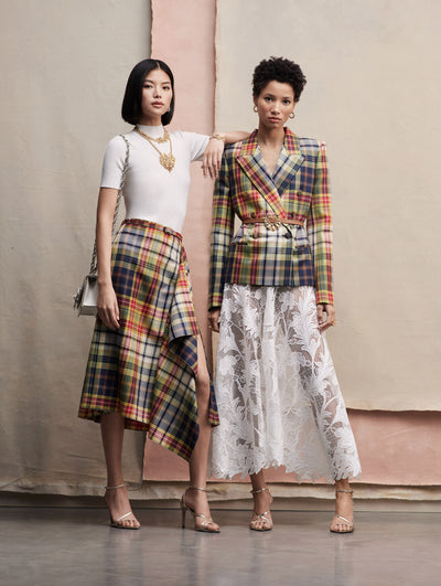 Two models stand next to each other. The model in the left is wearing a white mock-neck tee with a plaid skirt with a slit and ruffle detail. The model in the right wears a plaid blazer with a lace skirt under.