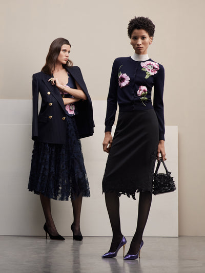 Two models pose together. The model in the left wears a navy lace skirt, with a navy tank printed with pink poppies, and a navy pinstripe blazer over. The model in the right wears a navy cardigan with pink poppies, paired with a black midi skirt.