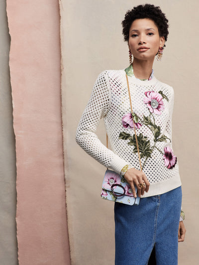 The model wears a white crochet pullover with an embroidered poppy, paired with a denim maxi skirt and the O Pochette bag with poppies.