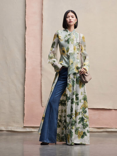 The model poses in a Light blue long blouse with yellow flowers printed, the blouse is tied around the neck and is opened from the waist down. It is paired with some blue jeans and a beige handbag. Front view