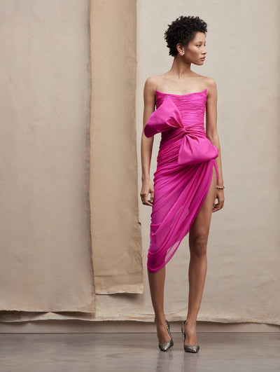 The model poses in a hot pink mini dress with a big bow in the side and slit, paired with pointy silver heels. Front view.