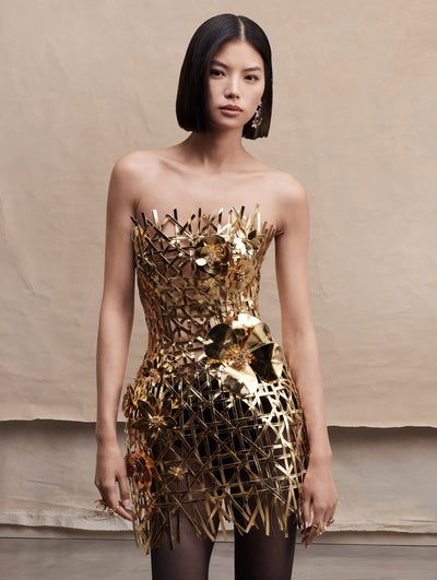 Model in strapless mini gold woven dress with three-dimensional flowers across.