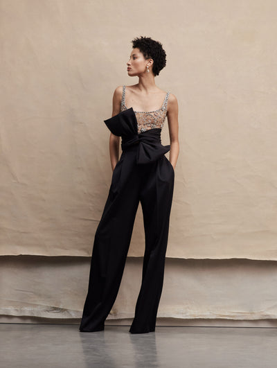 The model poses in a jumpsuit that has an embellished top, and black pants with a big bow around the waist. Front View
