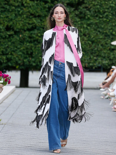 The model is wearing a long white coat with black fringe, a pink high neck silk blouse and dark blue denim pants. 
