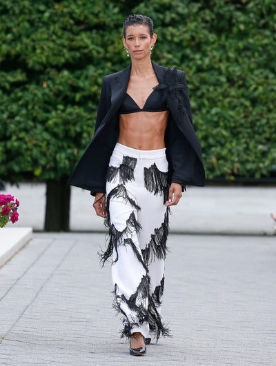 The model is wearing a black blazer with a flower on the lapel, a black bra and white suit pants with black fringe tassels. 