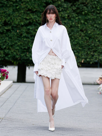 The model is wearing a white long sleeve shirt dress with a mini floral skirt. 