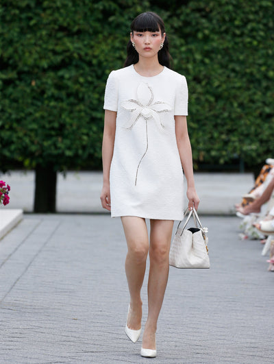The model is wearing a short sleeve white textured guipure mini dress adorned with a large orchid threadwork appliqué.