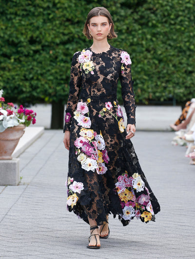 The model is wearing a long sleeve black lace dress with white, yellow and pink applique flowers. 