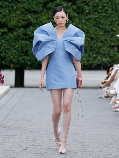 The model is wearing a blue mini dress with exaggerated bow sleeves. 
