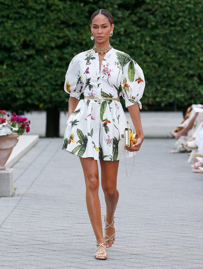A model is wearing a white dress with a botanical print featuring green leaves and colorful flowers, designed with voluminous puffed sleeves and a belted waist that flows into a short, pleated skirt. 