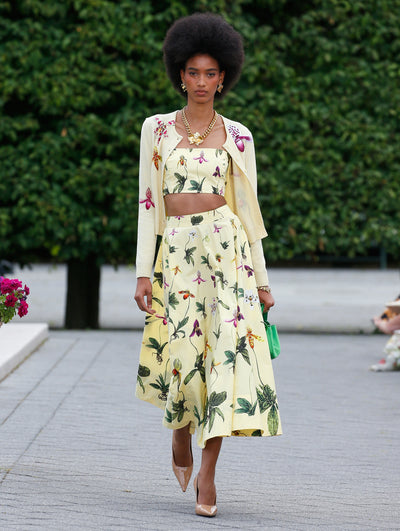 A model is wearing a coordinated outfit in a soft yellow color, consisting of a cropped top and a high-waisted, A-line midi skirt with a botanical print featuring green leaves and colorful flowers. She pairs the look with a matching cardigan.