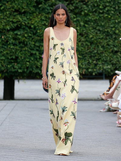 A model is wearing a sleeveless, floor-length dress in a soft yellow color, adorned with delicate floral embroidery featuring various green leaves and vibrant flowers. 