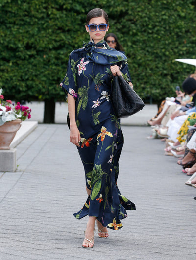 A model is wearing a flowing navy dress with a botanical print featuring vibrant green leaves and colorful flowers. The dress has a high neckline tied with a bow.