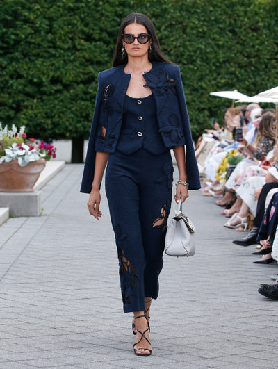 A model is wearing a navy three-piece set consisting of a fitted tank top, tailored trousers, and a cropped jacket adorned with floral cut-out details. 