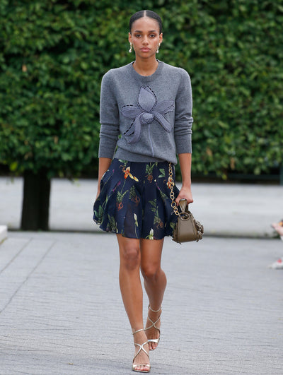 A model is wearing a gray sweater with a large embroidered floral design on the front, paired with a navy skirt featuring a delicate botanical print. 