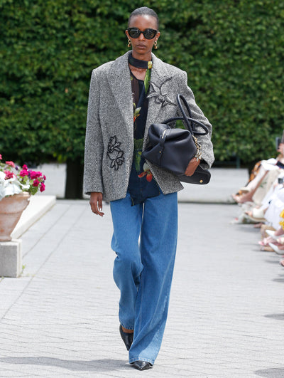 A model is wearing a gray herringbone blazer adorned with black floral appliqués, paired with a navy blouse featuring a floral pattern and wide-leg blue jeans. 
