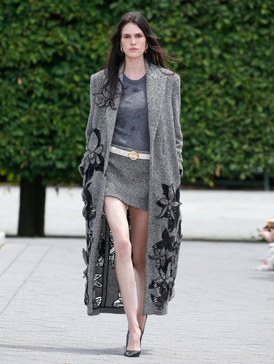 A model is wearing a long, gray herringbone coat with large black floral appliqués, paired with a matching gray mini skirt and a textured sweater.